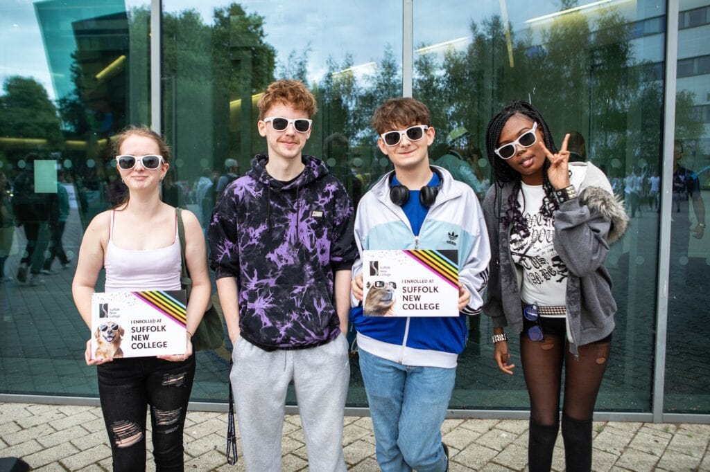 Four learners wearing sunglasses outside the Ipswich campus building holding enrolment signs.
