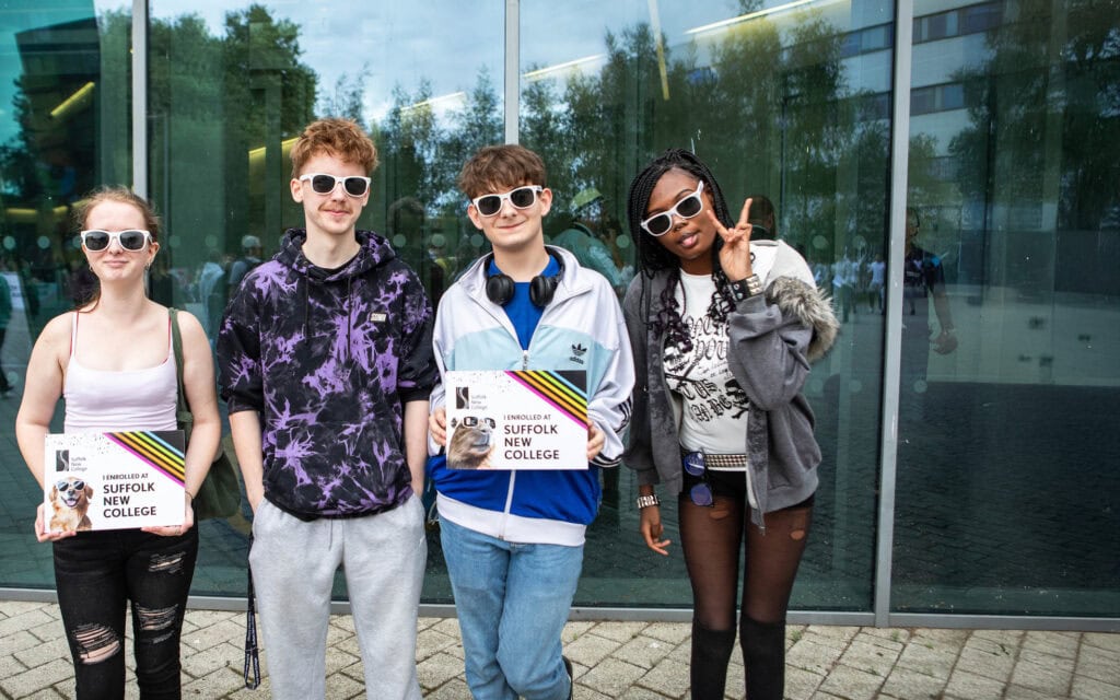 Four learners wearing sunglasses outside the Ipswich campus building holding enrolment signs.