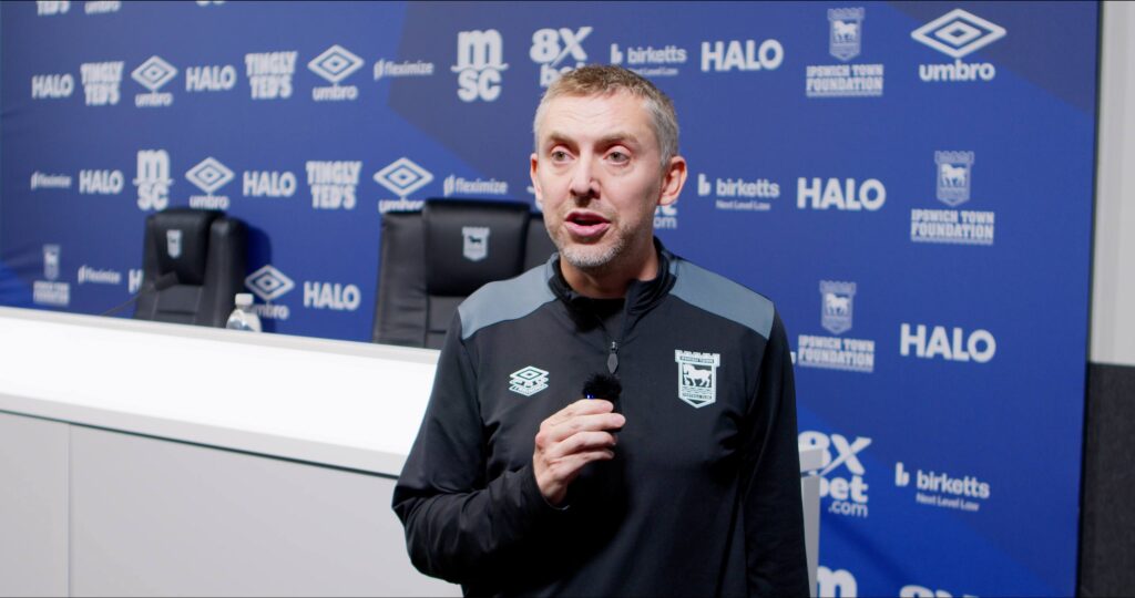 Professional man giving a presentation at Suffolk New College sports event, wearing sportswear with college logo, in front of a branded backdrop, focusing on sports education and college achievements.