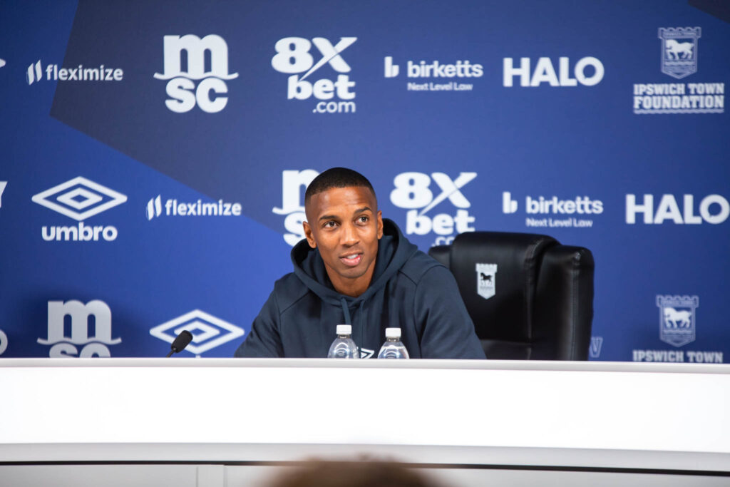 Bright smiling man at a sports press conference, sitting at a table with water bottles, in front of a blue backdrop with sponsor logos.
