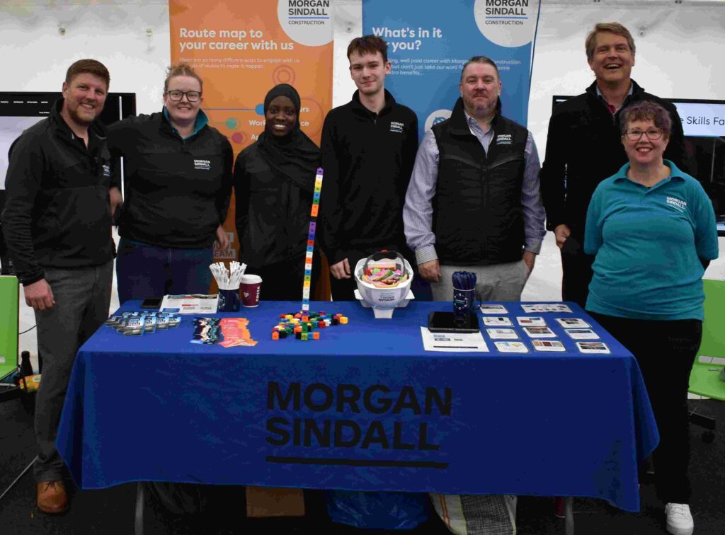 Diverse group of students and staff at Suffolk New College standing behind an informational booth, promoting careers and courses with Morgan Sindall banners in the background.
