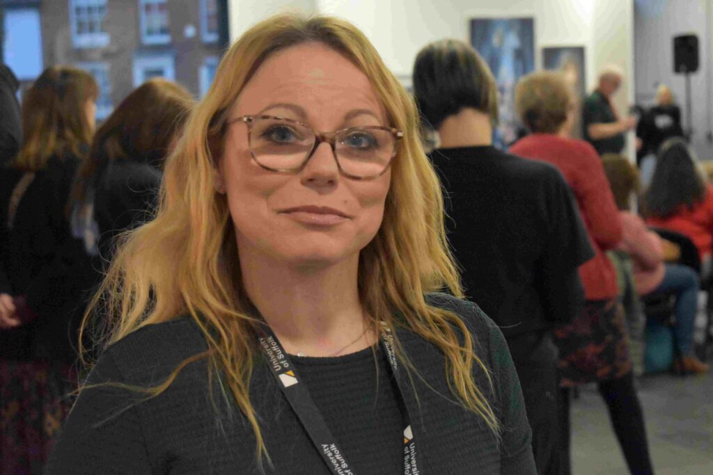 A confident female student with glasses at Suffolk New College, surrounded by diverse students in a lively classroom environment, showcasing education and community focus.
