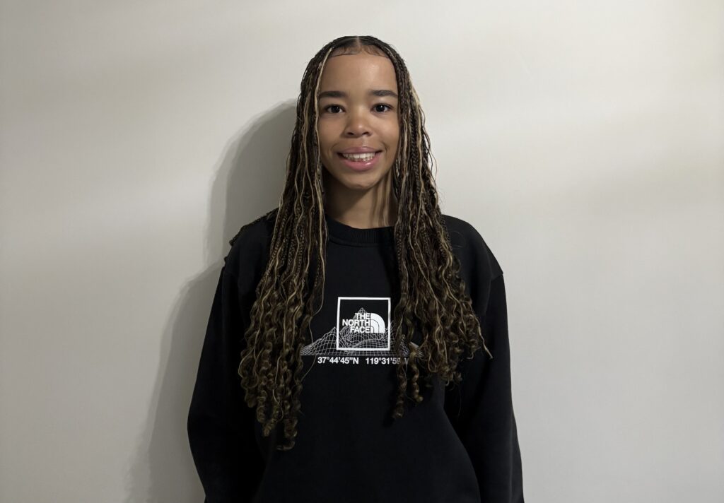 A young woman with long, styled braids attire, smiling confidently against a plain wall, showcasing Suffolk New College's vibrant student community and diverse education opportunities.