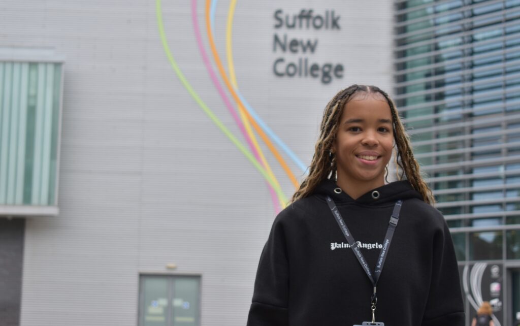 A smiling student wearing a black hoodie with Suffolk New College signage standing in front of the modern college building, showcasing advanced facilities and vibrant student life.