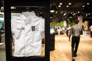A chef's white uniform featuring Suffolk New College branding displayed at a college event, with students and staff in a vibrant dining area in the background.