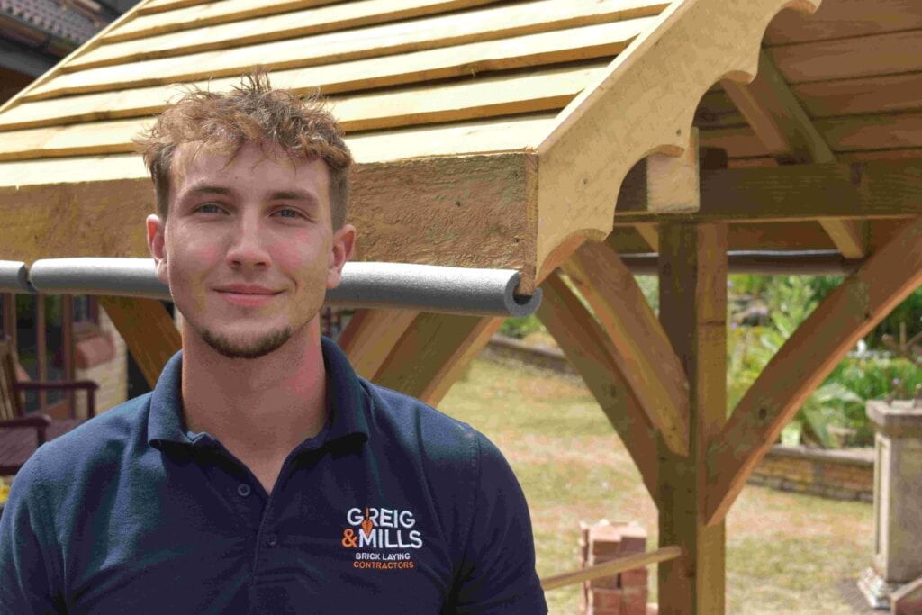 Young man in navy polo shirt with "Greig & Mills Brick Laying Contractors" logo, standing outdoors near a wooden garden structure.