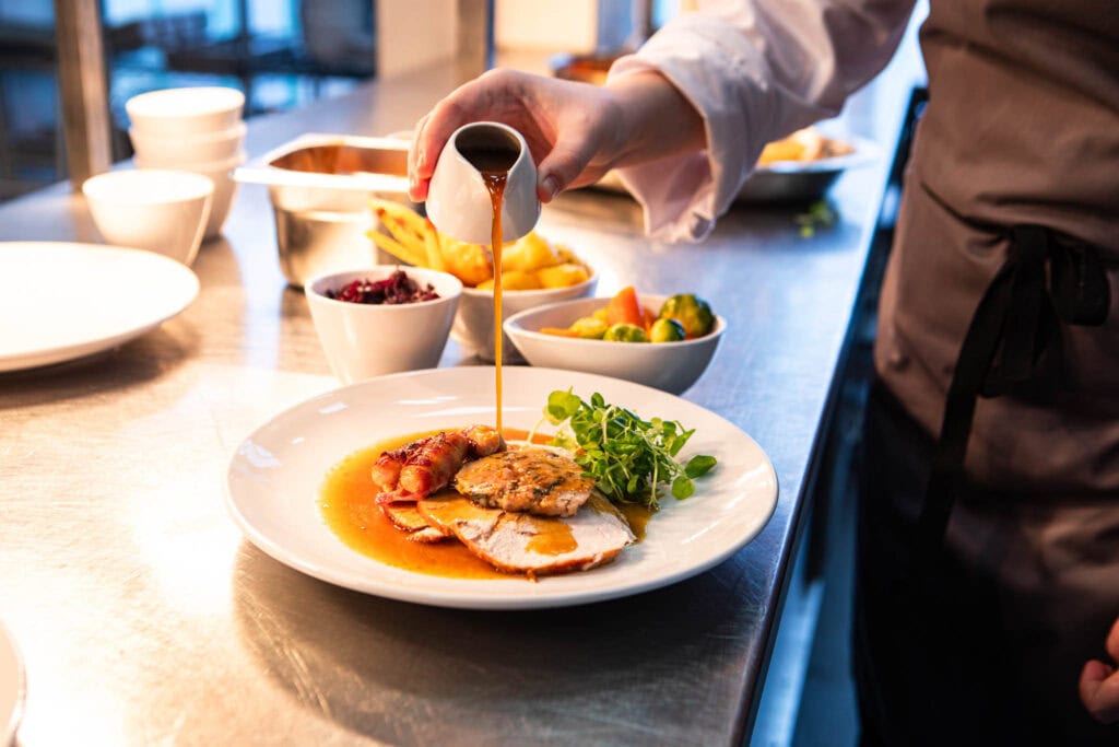 A student chef in culinary arts pouring sauce over a plated gourmet meal in a modern college kitchen.