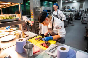 A group of students in chef uniforms preparing meat and learning culinary skills in a modern college kitchen.