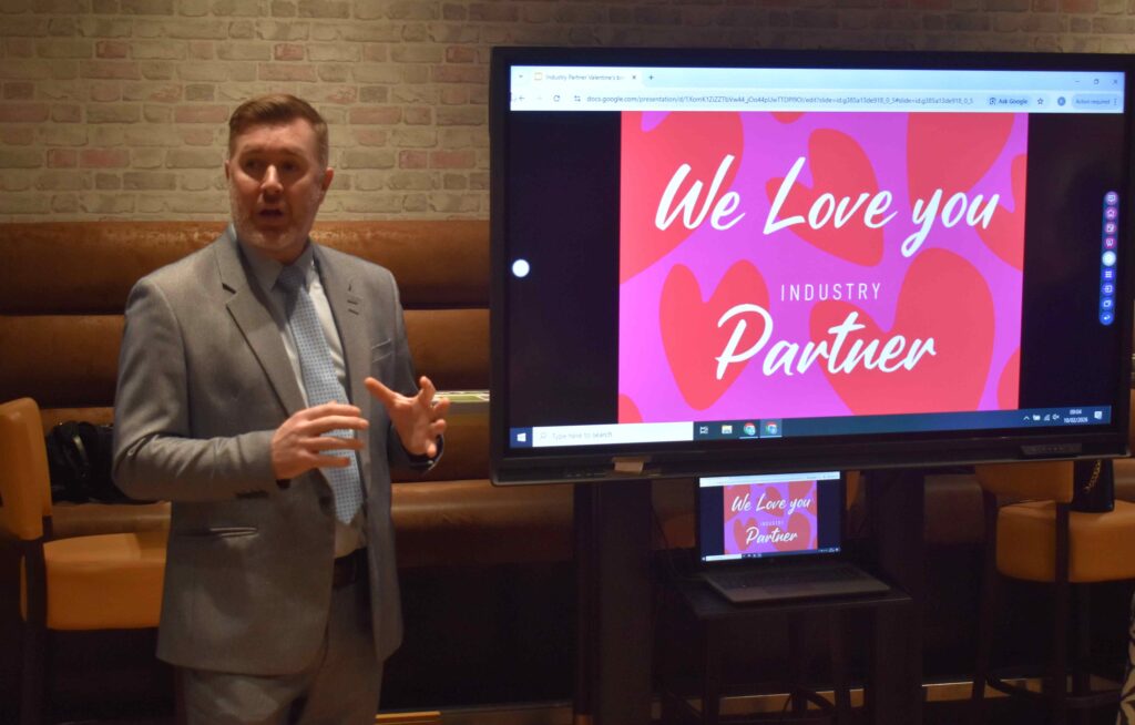 A man in a grey suit presenting at Suffolk New College's Valentine’s themed event, showcasing industry partnerships and apprenticeships to students and partners.