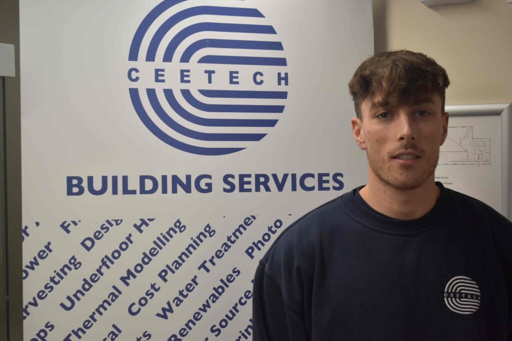 A young man in a navy sweatshirt standing in front of a Suffolk-based building services company banner, highlighting electrical and mechanical apprenticeships at Suffolk New College.