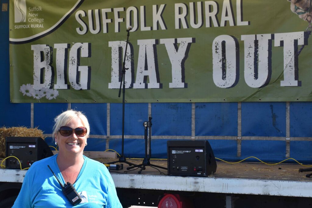 A smiling woman wearing sunglasses and a blue shirt at the Suffolk Rural Big Day Out event, with a stage and banner in the background promoting family fun and community activities.