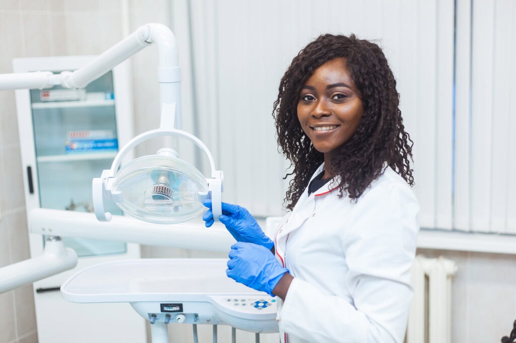 A smiling dental nursing student in a white coat and blue gloves working with dental equipment in a clinical training environment at Suffolk New College.
