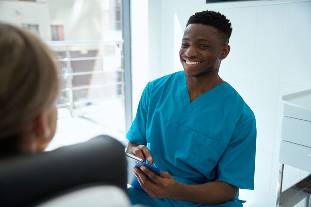 A smiling dental nursing student in blue scrubs engaging with a patient in a clinical setting at Suffolk New College, highlighting dental care training and patient interaction.