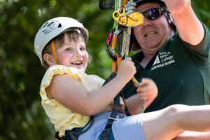 A young girl enjoying a zip-lining adventure during the Big Day Out family fun day at Suffolk New College, with an instructor assisting her in a lush outdoor setting.