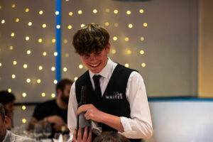 Young chef in white uniform and black apron smiling while presenting a dish at Suffolk New College's Chefs' Whites Restaurant, showcasing culinary skills and hospitality training.