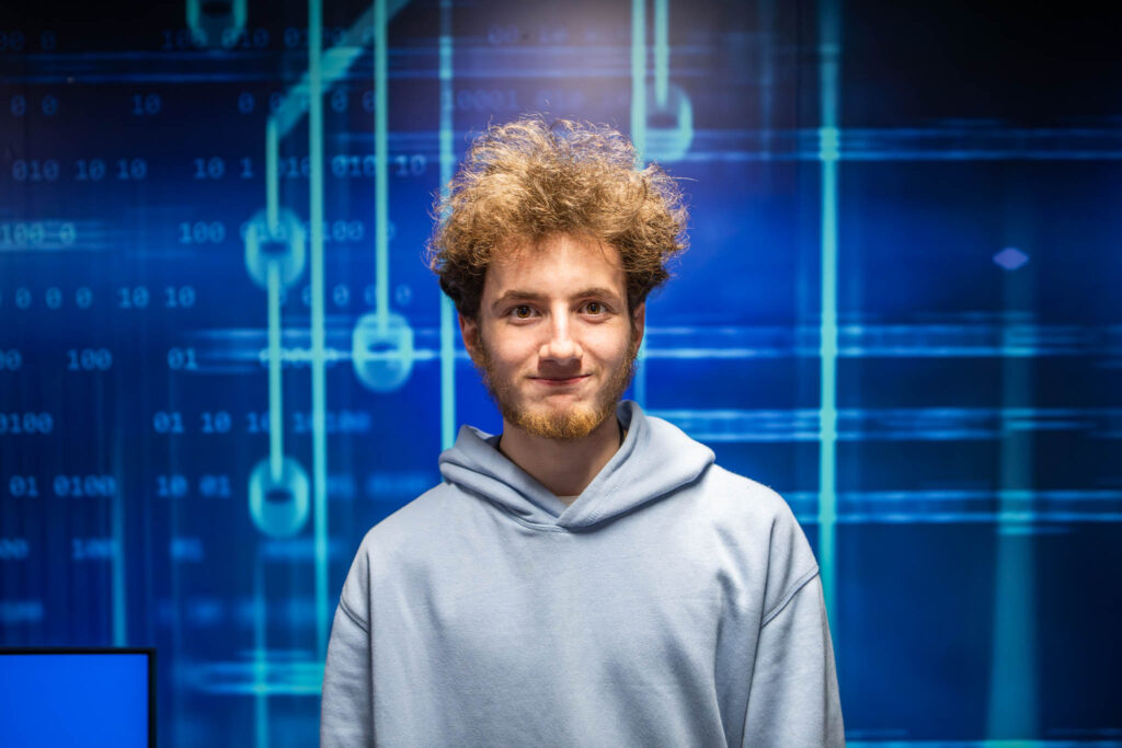 A young man with curly hair and a hoodie standing in front of a digital gaming and data background, representing Suffolk New College's esports and digital media courses.