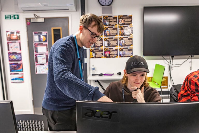 A teacher assisting a student in a game design class at Suffolk New College, with computers and educational posters in the background.