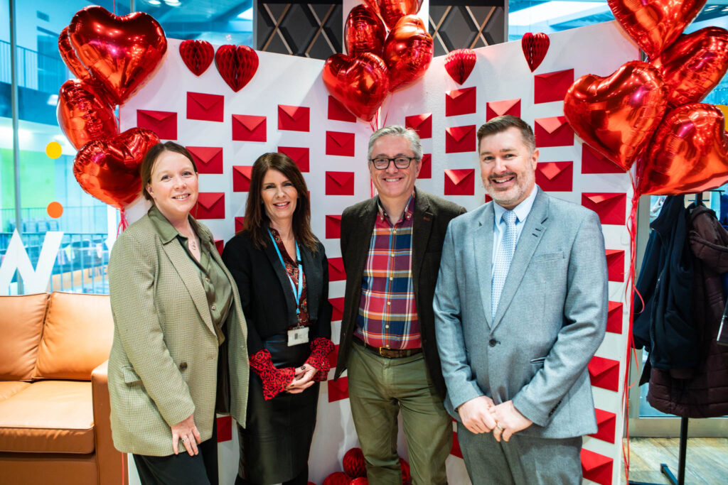 Group of four professionals at Suffolk New College during a Valentine's themed event, with heart-shaped balloons and red envelopes on the wall, promoting apprenticeships and industry partnerships.