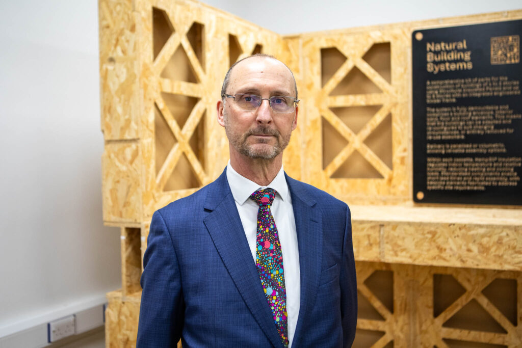An image of a man in a blue suit and colorful tie standing in front of a wooden construction display at Suffolk New College, highlighting civil engineering training and apprenticeships.