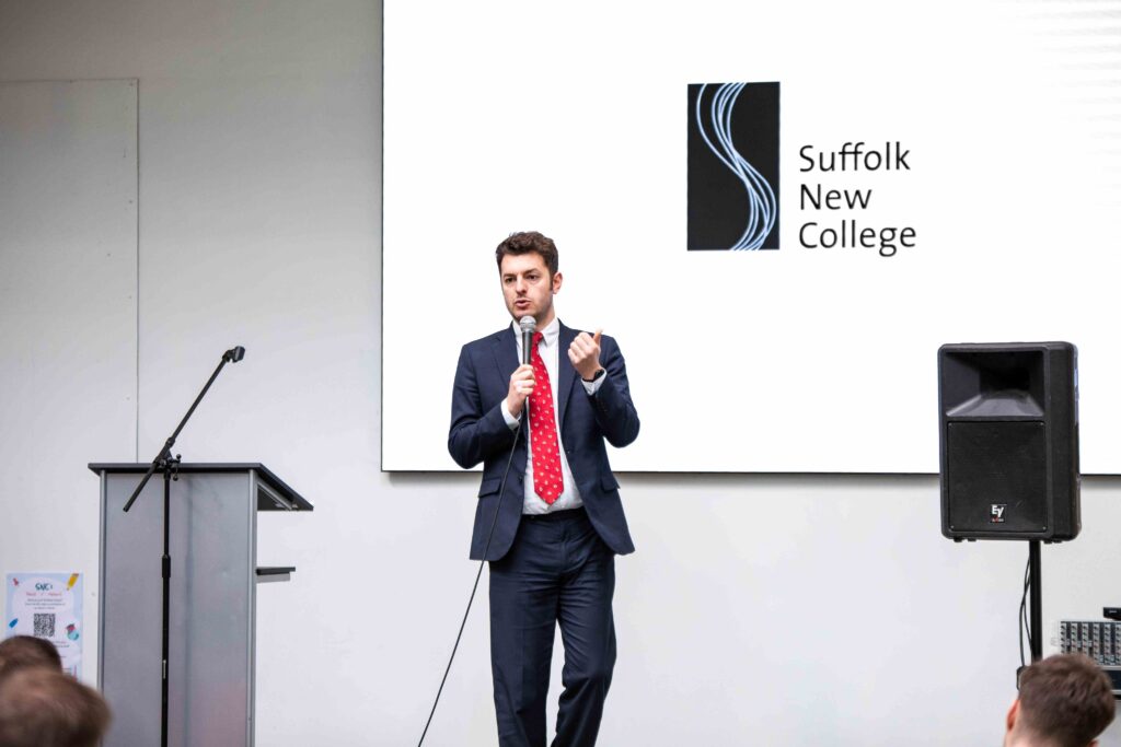 A Suffolk New College student giving a speech during a presentation, with the college logo displayed on the screen behind him, in a classroom setting.