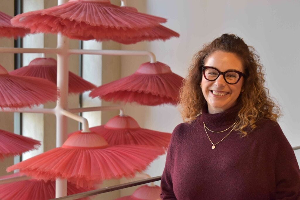 A smiling woman with curly hair and glasses standing in front of an art installation featuring pink umbrellas, highlighting Suffolk New College's creative arts programs.