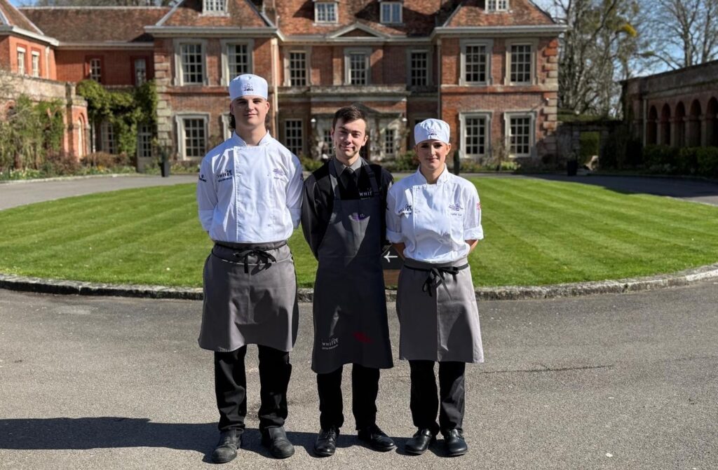 Three young restaurant team finalists in chef uniforms standing outdoors in front of a historic building, representing Suffolk New College's culinary excellence and teamwork.