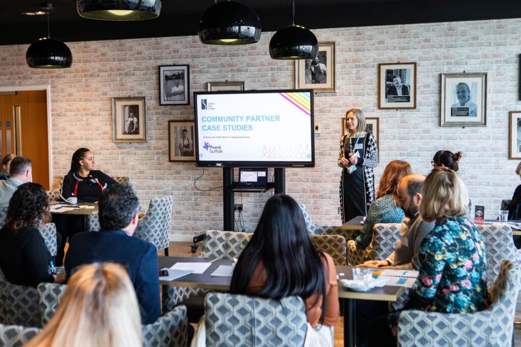 A speaker presents at Suffolk New College during a community partnership event, with attendees listening attentively in a modern, well-lit room decorated with framed photos.