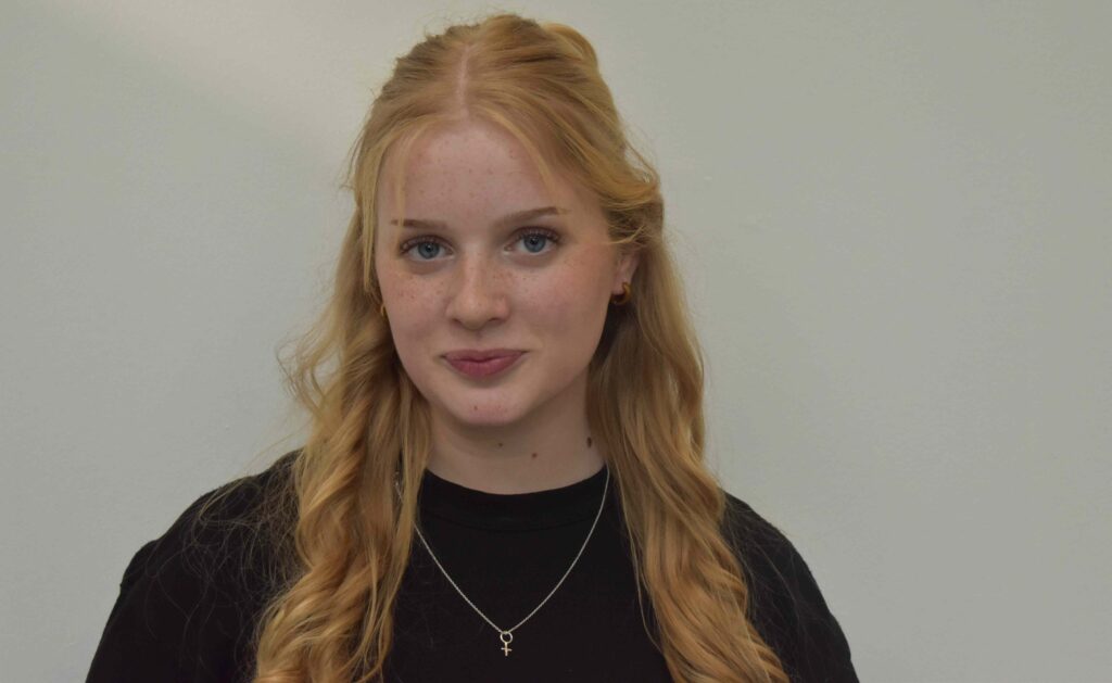 A young woman with long red hair and freckles, wearing a black top and silver necklace, standing against a plain background, representing Suffolk New College students.