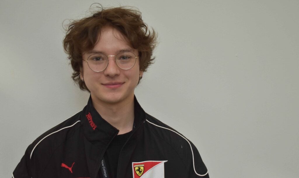 A young student with curly hair and glasses, wearing a black sports jacket with a Ferrari logo, standing against a plain light background.