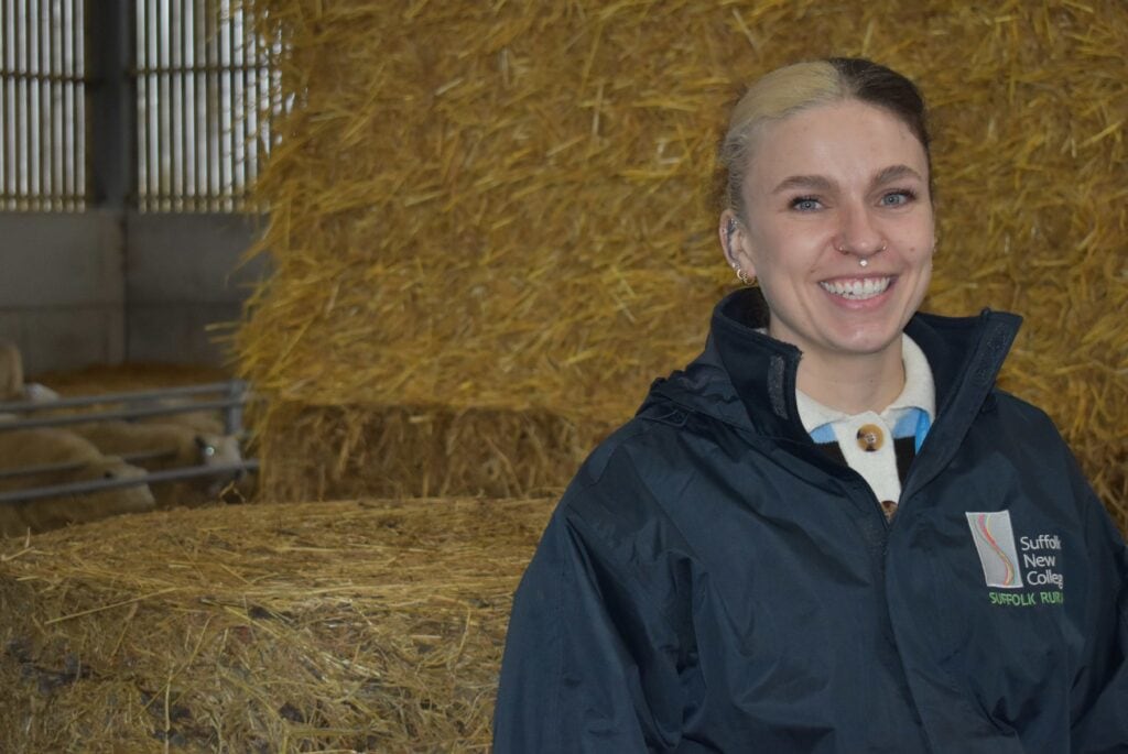 A smiling female student at Suffolk New College, dressed in a navy jacket, standing inside a farm building with hay bales, highlighting increased female participation in rural farming courses.