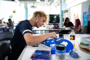 A student working on an art project in a bright, modern classroom at Suffolk New College, focusing on creative art and design techniques with various art supplies around.