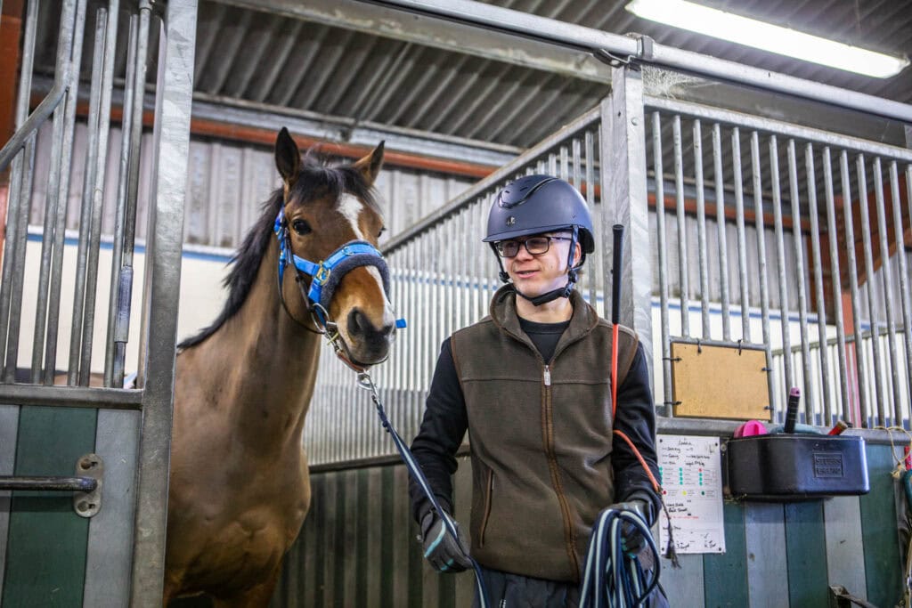 Student in riding helmet and jacket with a horse in a stable, demonstrating equine care and handling skills at Suffolk New College.