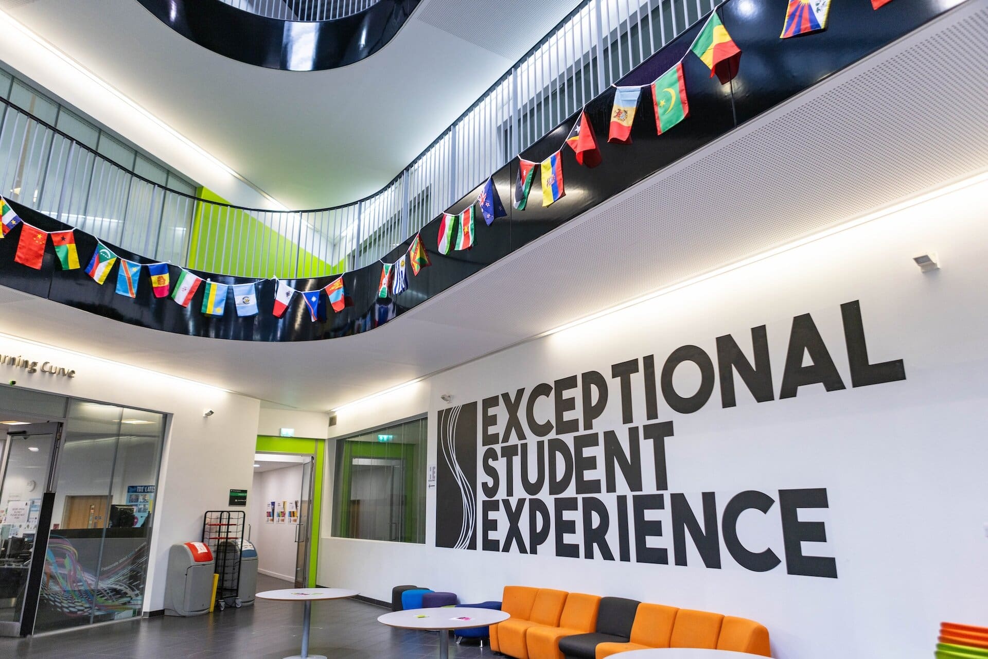 Interior view of Suffolk New College showcasing a welcoming lounge area with vibrant seating, international flags, and a prominent "Exceptional Student Experience" sign, emphasizing inclusivity and su.