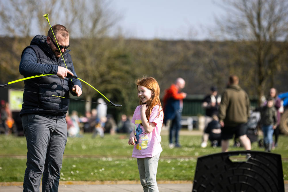 A man and a young girl enjoying a fun outdoor activity at Suffolk New College's spring family event, with other families and children in the background.