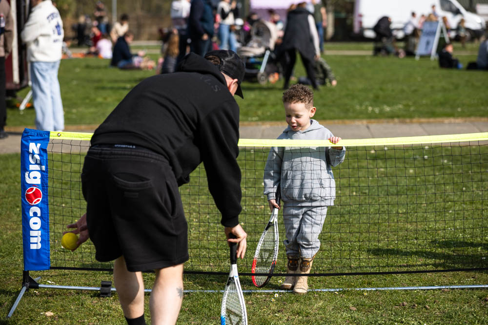 A young boy and an adult playing tennis at the Suffolk New College spring family fun day, with other visitors enjoying outdoor activities in the background.