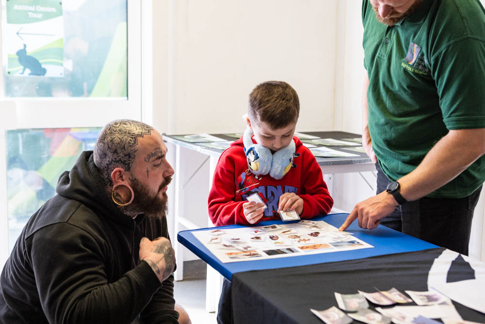 A young boy wearing a face mask and red hoodie engaging with an adult at Suffolk New College during a spring family fun day event, with informational displays on the table.