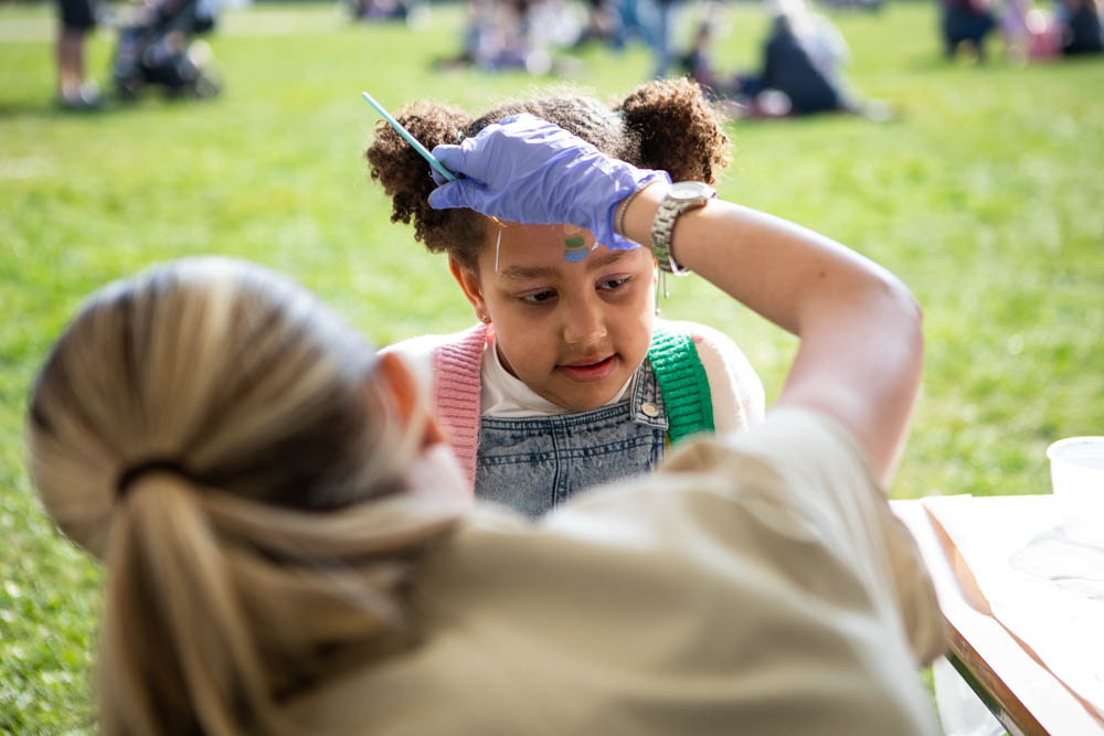 A young girl receives a fun spring-themed face painting at Suffolk New College's family event, with other families enjoying outdoor activities in the background.