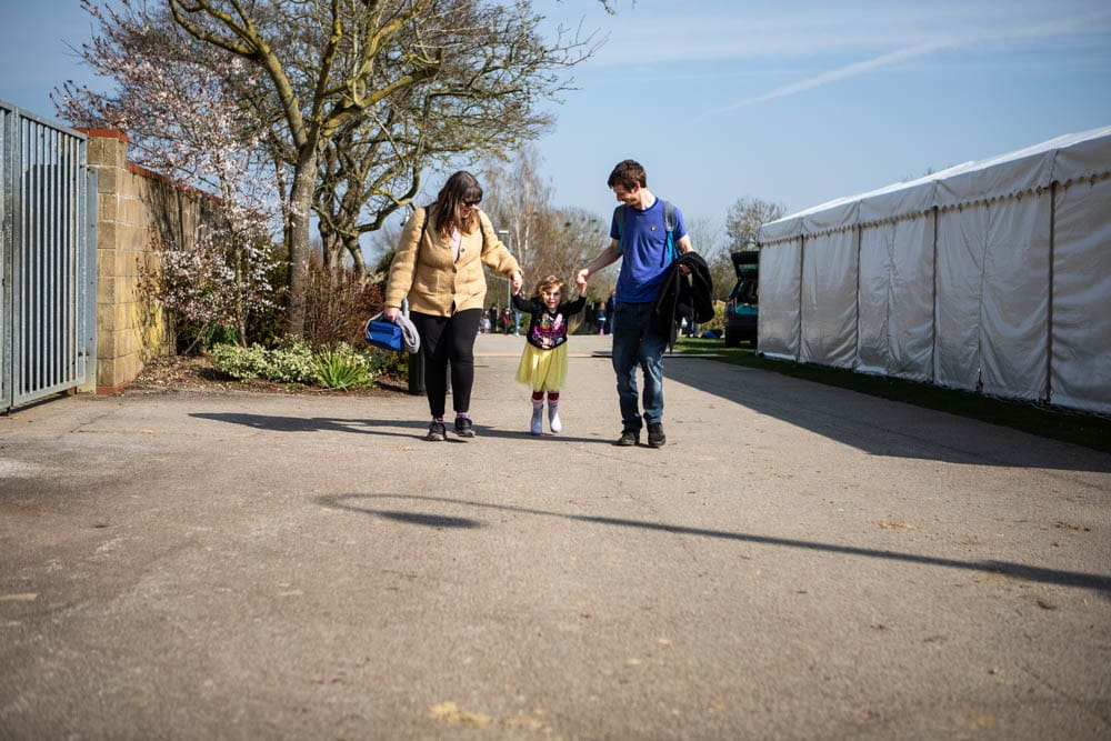 An outdoor scene of a family walking together at Suffolk New College's spring family fun day, featuring children and parents enjoying a community event with tents and blooming trees.