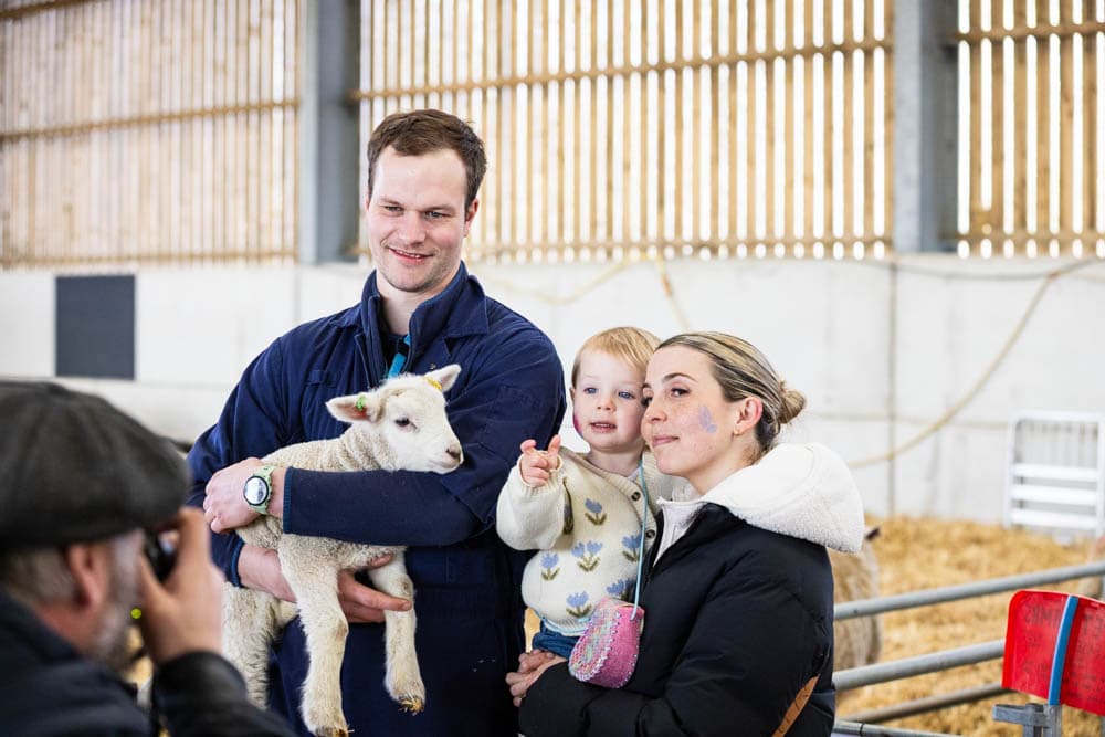Family enjoying a lamb at Suffolk New College's spring fun day, with children and adults engaging in farm activities during the 'lambtastic' event.