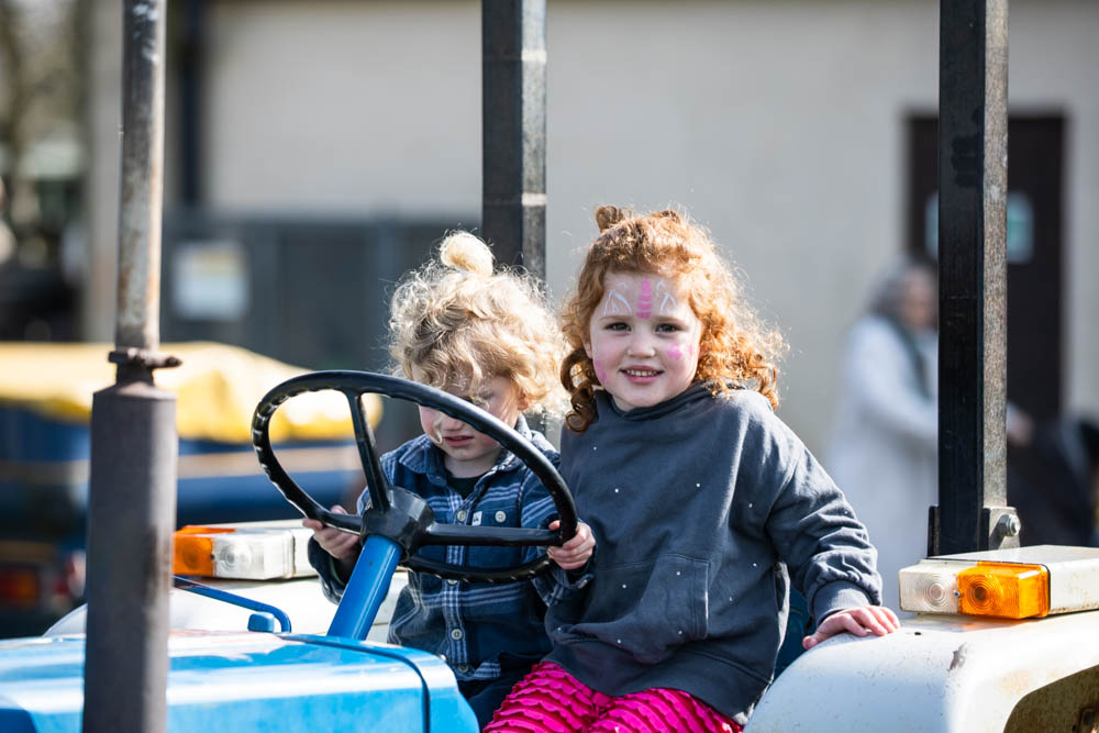 Two young girls enjoying a tractor ride at Suffolk New College's spring family fun day, part of the 'lambtastic' event attracting families and children for outdoor activities.