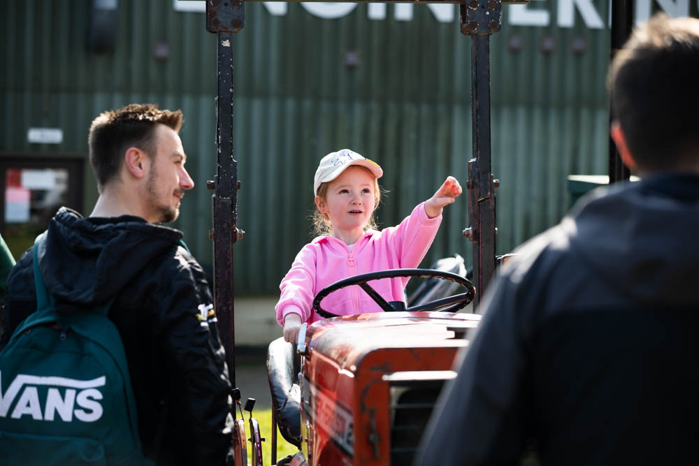 Young girl in pink jacket and white cap enjoying tractor ride at Suffolk New College's spring family fun day, attracting families for outdoor activities and entertainment.