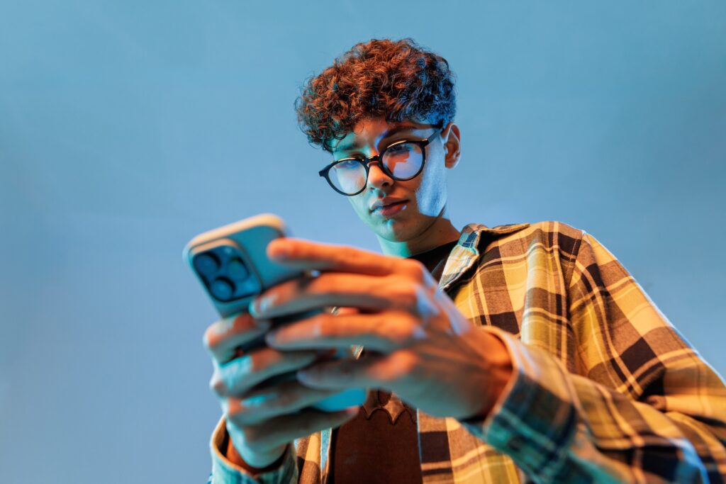 Young student with curly hair and glasses engaging with social media on a smartphone, highlighting emerging social media trends among students at Suffolk New College.