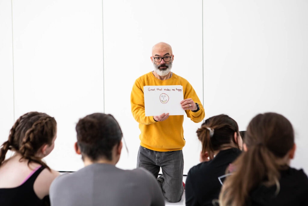 A college instructor holding a whiteboard with a smiley face, teaching a group of students in a modern classroom setting, promoting arts and creative skills development.
