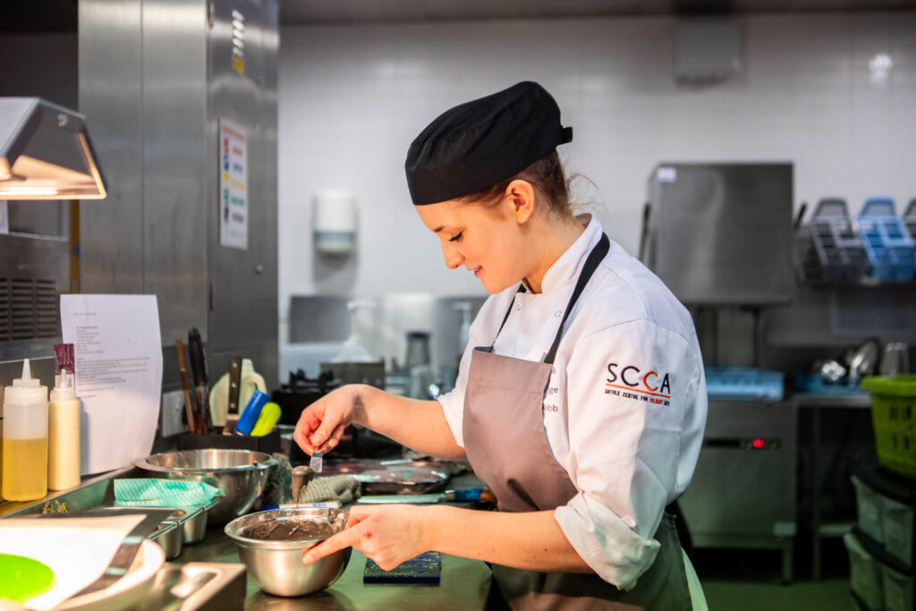 Young female culinary student at Suffolk New College wearing chef's hat and apron, preparing ingredients in a professional kitchen for a national restaurant competition.