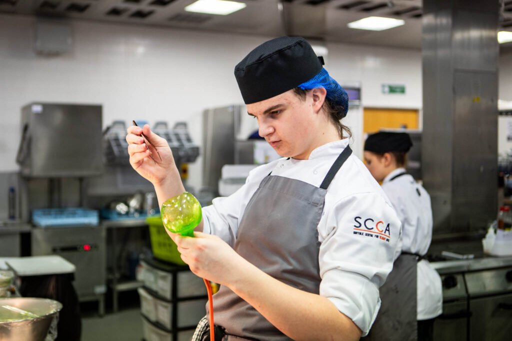 A young female culinary student at Suffolk New College carefully preparing a dish in the college's professional kitchen, demonstrating skills for a national restaurant competition.