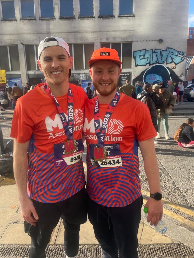 Two male runners in red Suffolk New College t-shirts and medals, smiling outdoors with a crowd and graffiti background.