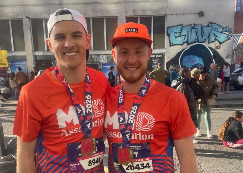 Two male runners in red Suffolk New College t-shirts and medals, smiling outdoors with a crowd and graffiti background.