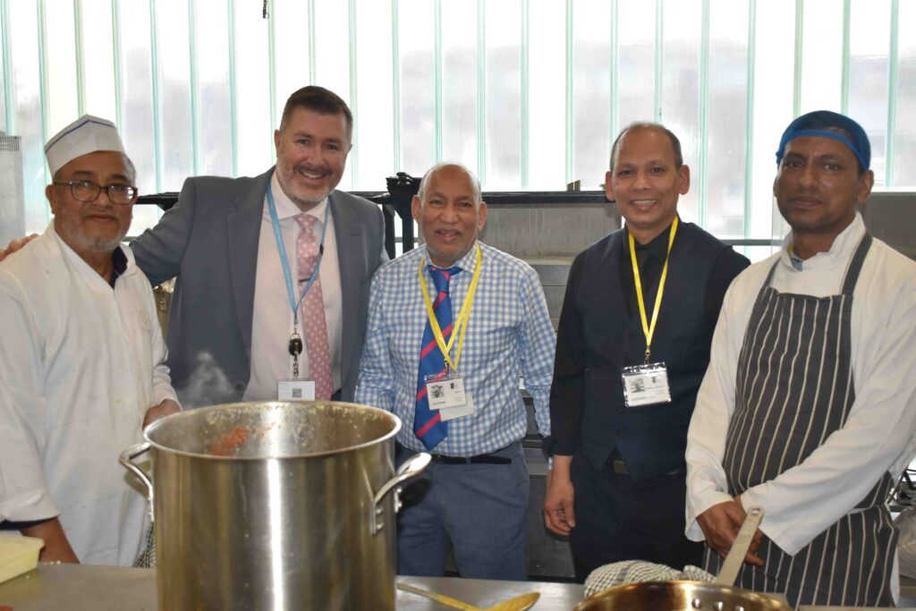 Group of five men, including chefs and culinary experts, posing in a kitchen with cooking utensils and a large pot, during a culinary masterclass at Suffolk New College.