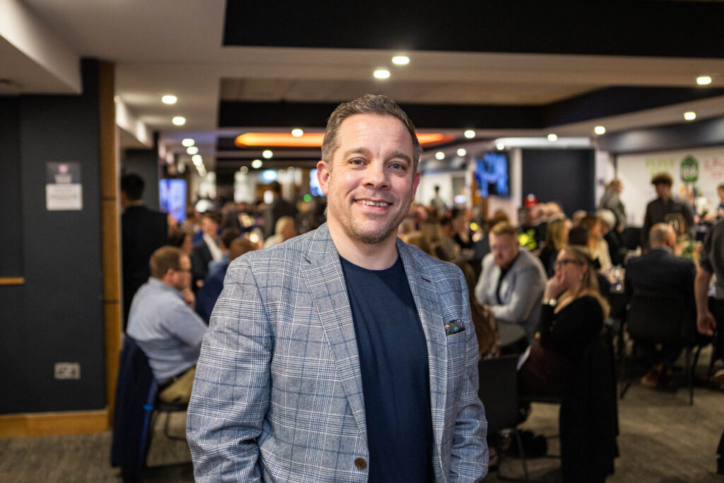 A college student attending the industry dinner at Ipswich Town Football Club, with a lively crowd of over 130 guests in the background, showcasing Suffolk New College's engagement with industry event.