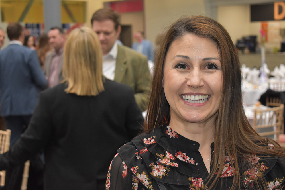 Smiling woman at Suffolk New College during the annual dinner celebrating reaching 200 industry partners, with attendees networking in the background.